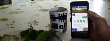 Hand holding a phone showing the iWalk Cornwall app